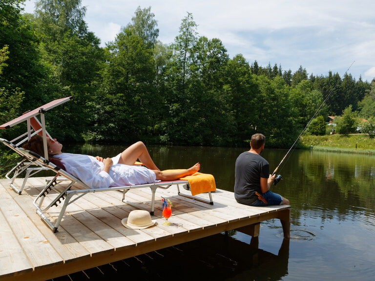 Frau entspannt auf Wellnessliege während ihr Freund im Naturweiher angelt im Hotel Deine Auszeit im Bayerischen Wald